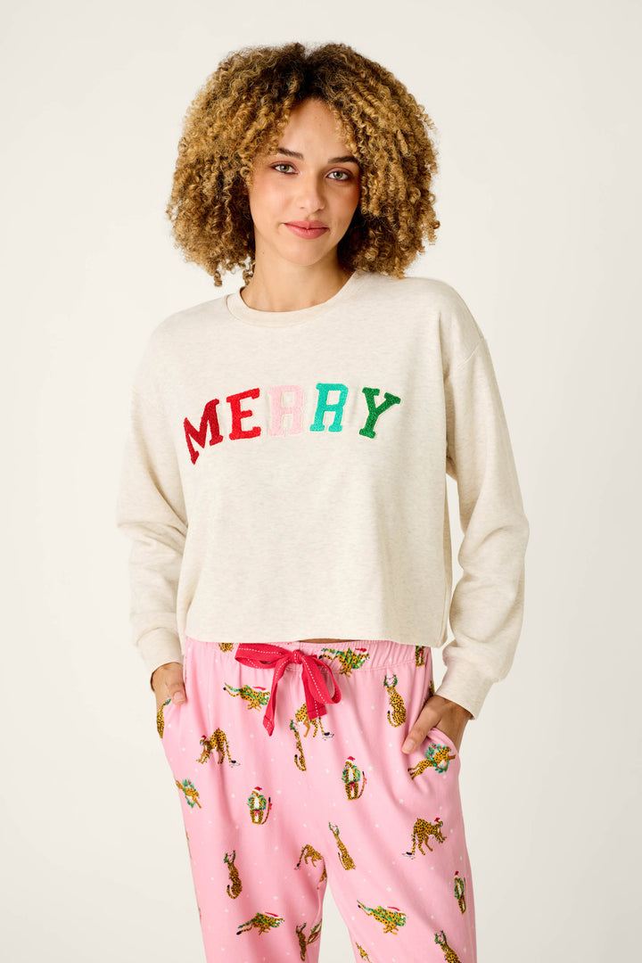 A person wearing a P.J. Salvage Very Merry Bouclé Merry Sweatshirt in cream with colorful, textured letters, paired with pink leopard-print pajama pants.