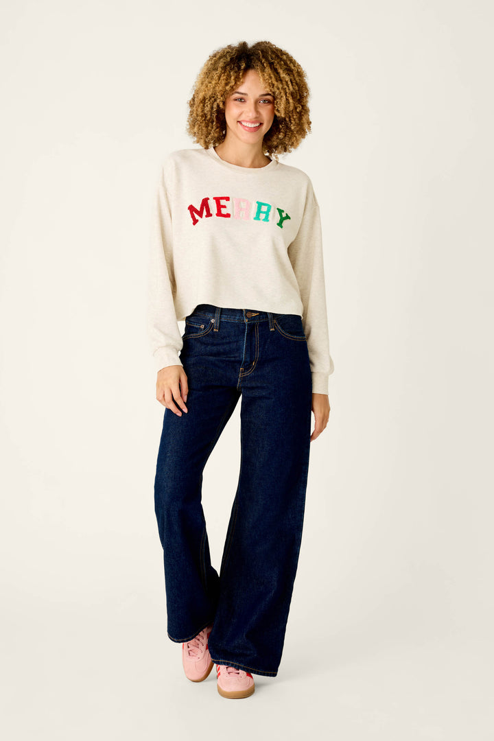 A smiling woman wearing a cozy cream-colored P.J. Salvage Very Merry Bouclé Merry Sweatshirt with MERRY in colorful letters, paired with dark blue wide-leg jeans and pink sneakers.