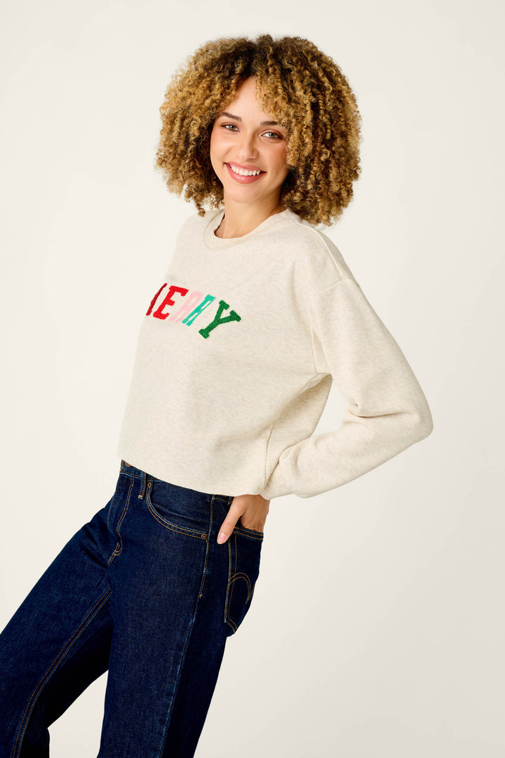 A smiling woman wears a cream P.J. Salvage Very Merry Bouclé Merry Sweatshirt with multi-colored, textured lettering across the chest and dark blue jeans.