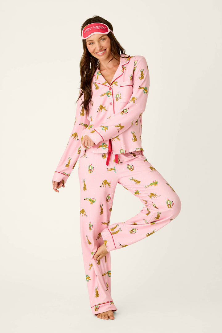 A smiling woman wearing a P.J. Salvage Very Merry Wild for the Holidays 3-Piece PJ Set featuring a Christmas cheetah print and a matching eye mask that says VERY MERRY.