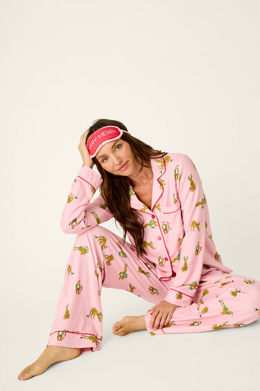 A woman wearing a P.J. Salvage Very Merry Wild for the Holidays 3-Piece PJ Set in leopard print with a matching sleep mask that reads Very Merry.