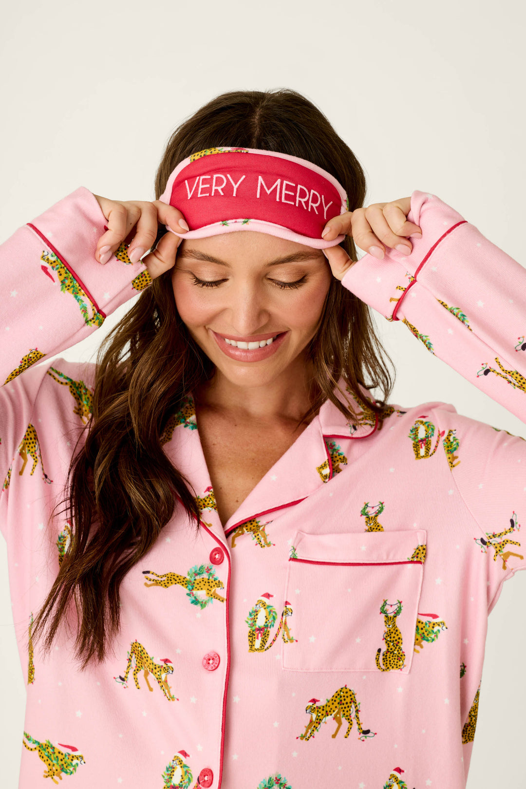 A woman wears the P.J. Salvage Very Merry Wild for the Holidays 3-Piece PJ Set, featuring pink, leopard-print, button-down pajamas with holiday wreaths and a matching sleep mask that reads VERY MERRY.