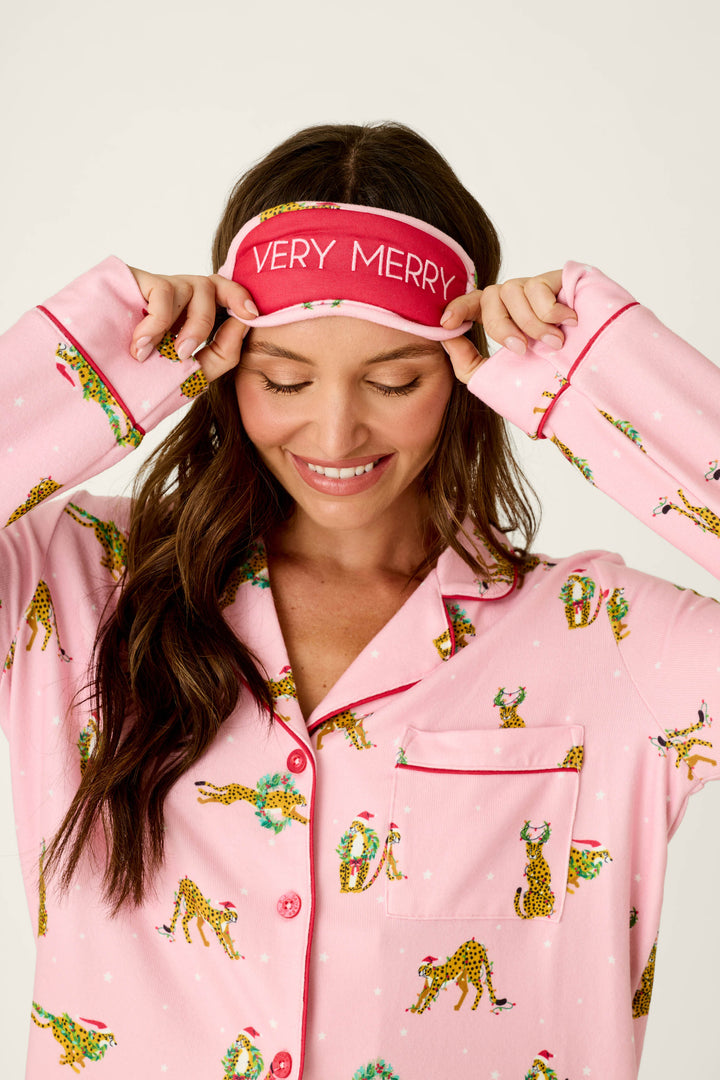 A woman wears the P.J. Salvage Very Merry Wild for the Holidays 3-Piece PJ Set, featuring pink, leopard-print, button-down pajamas with holiday wreaths and a matching sleep mask that reads VERY MERRY.