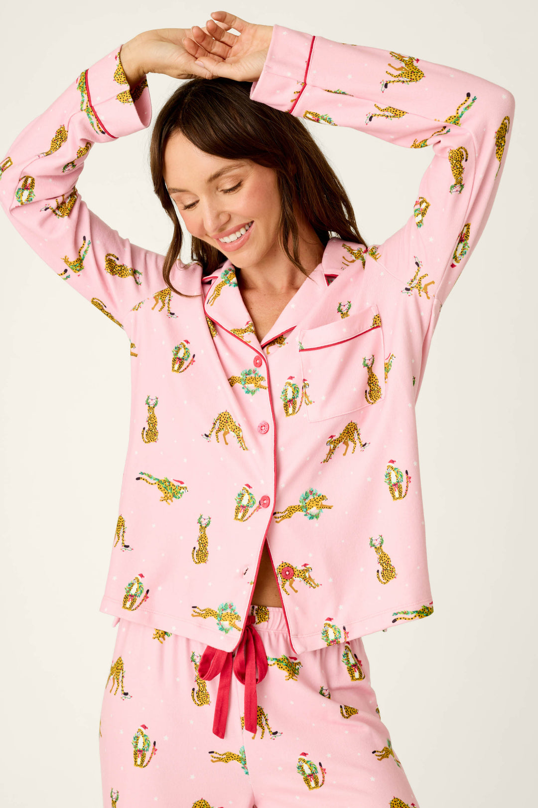 A person wearing the Very Merry Wild for the Holidays 3-Piece PJ Set, featuring a pink pattern of cheetahs in Santa hats and festive wreaths.