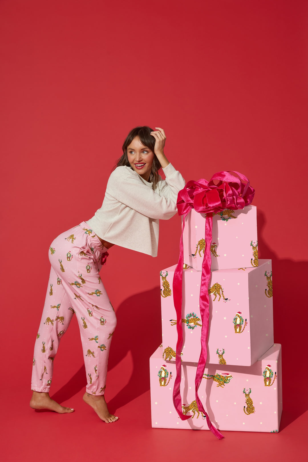 A smiling woman wearing a beige sweater and P.J. Salvage Very Merry Wild for the Holidays Jogger Pants leans against a stack of three gift boxes wrapped in matching paper with a large pink bow.