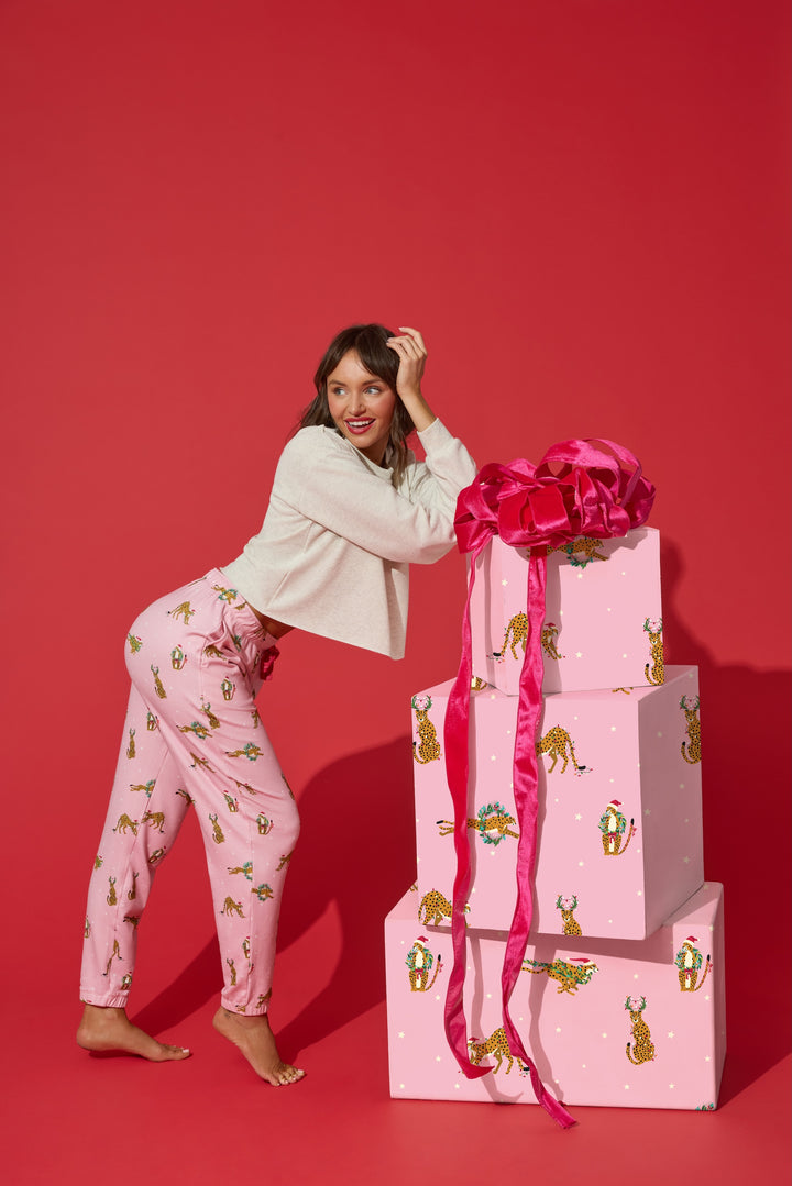A smiling woman wearing a beige sweater and P.J. Salvage Very Merry Wild for the Holidays Jogger Pants leans against a stack of three gift boxes wrapped in matching paper with a large pink bow.