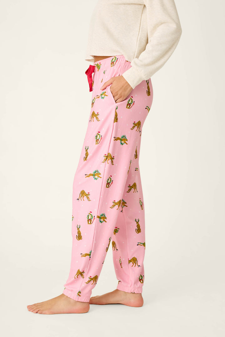 A side view of a person in pink P.J. Salvage Very Merry Wild for the Holidays Jogger Pants featuring a repeating pattern of cheetahs in Christmas hats and scarves, paired with a white long-sleeved shirt.