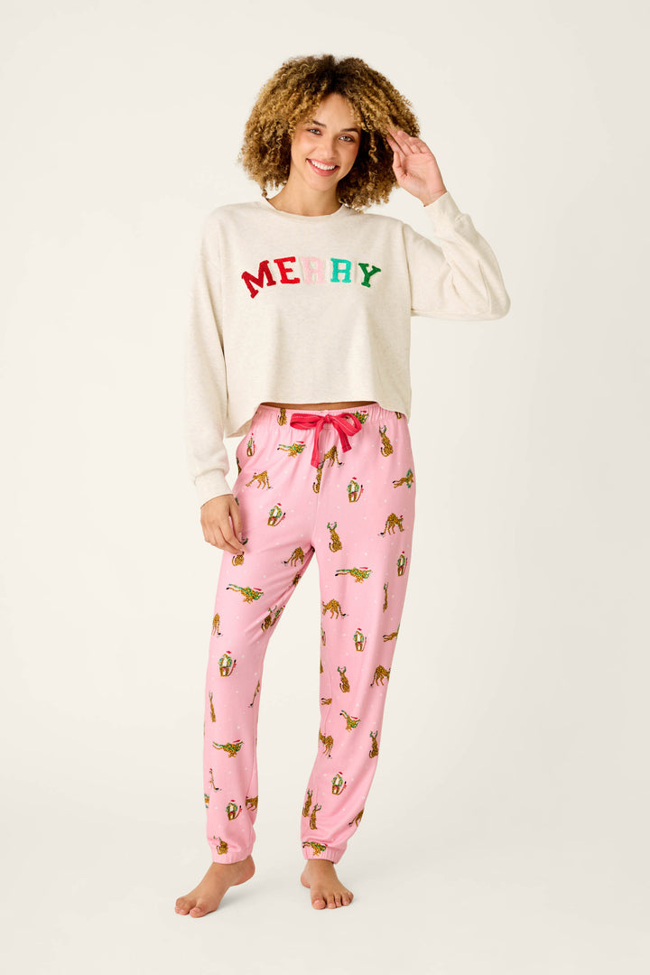 A person wearing a cream-colored MERRY sweatshirt and P.J. Salvage Very Merry Wild for the Holidays Jogger Pants in a pink butter jersey leopard print with a pattern of animals wearing holiday hats.
