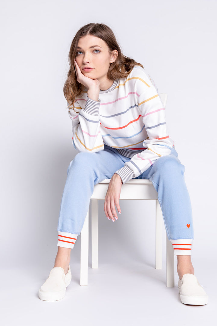 Woman sitting on a white stool wearing a colorful striped sweater and light blue pants with red accents.
