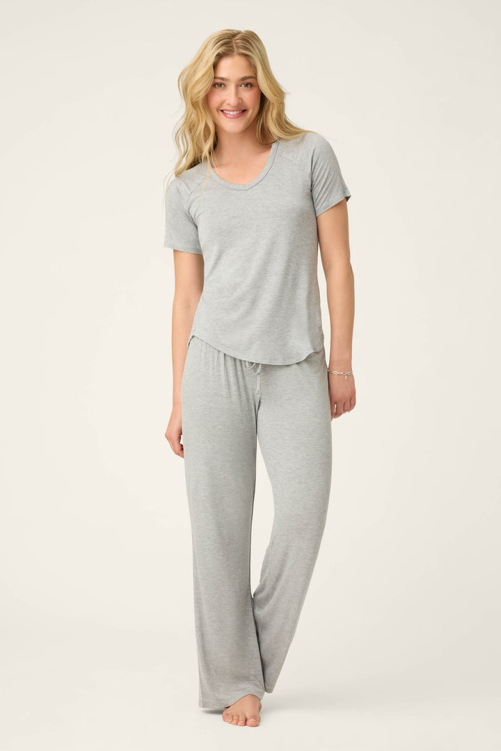 A person with long, wavy blond hair smiles barefoot on a light background, wearing the P.J. Salvage Modal Basics Heather Grey PJ Set, featuring a matching V-neck tee and tie-waist pant in soft jersey.
