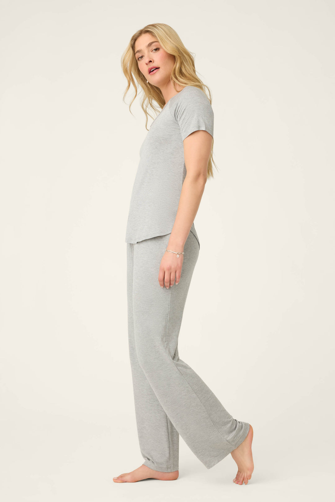 A woman with long blonde hair stands barefoot in profile against a plain background, wearing the P.J. Salvage Modal Basics Heather Grey PJ Set featuring a V-neck tee and matching loose tie-waist pant.