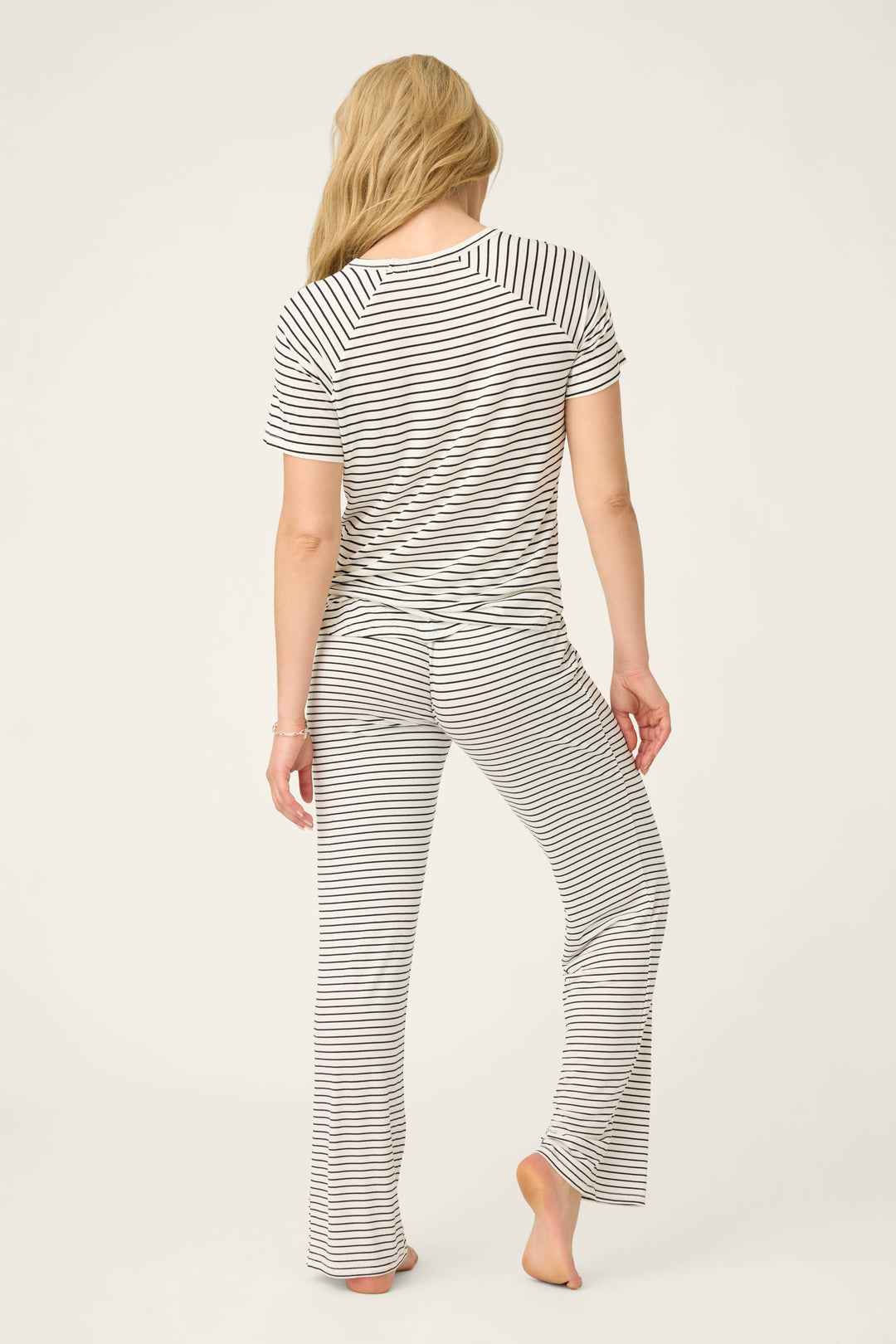 A person with long blonde hair stands barefoot, facing away, wearing the P.J. Salvage Modal Basics Striped PJ Set— a black-and-white striped V-neck tee and tie-waist pant— against a plain light background.