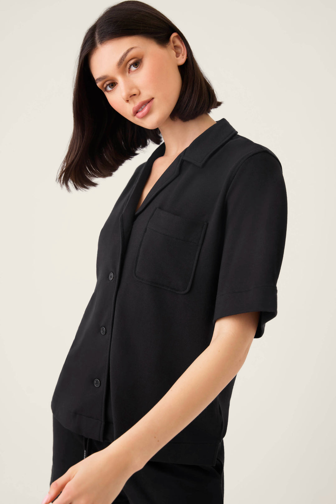 A woman with straight dark hair models the P.J. Salvage Jammie Essentials Short Black PJ Set, featuring a black notch collar top. She stands against a plain light background, looking at the camera.