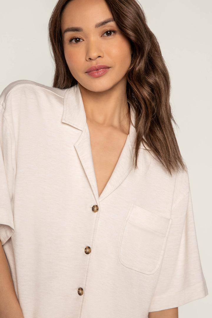 A person with long brown hair wears the P.J. Salvage Jammie Essentials Short Oatmeal PJ Set, featuring a cream-colored notch collar top with short sleeves and a front pocket, standing against a plain light background.