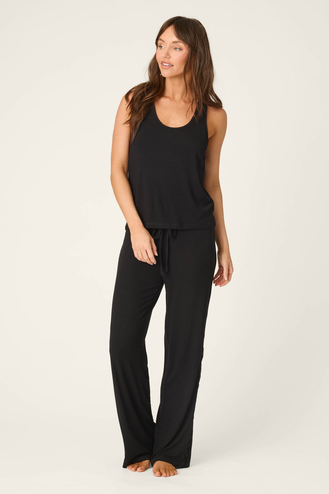 A woman stands barefoot against a plain background, wearing the P.J. Salvage Lacey Basics Black Lounge Set. She gazes left with one hand by her side, showcasing the sleeveless lace-trimmed tank and matching modal pants.