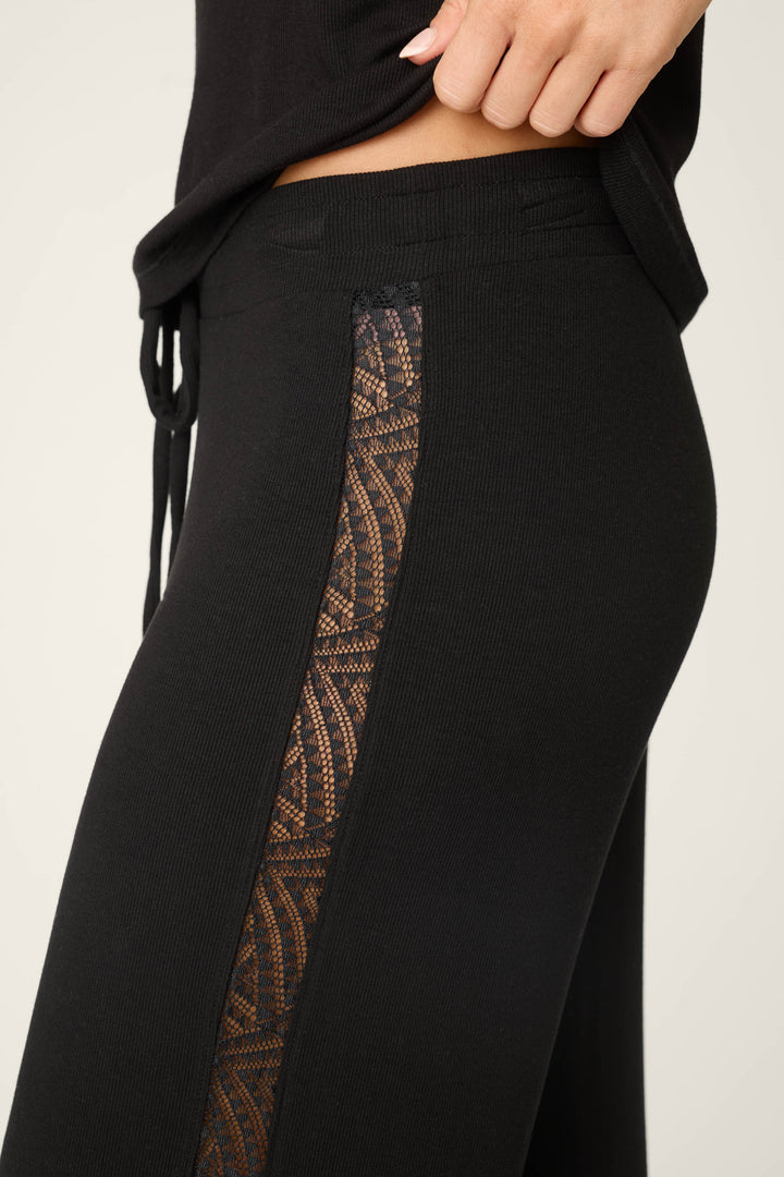 A person models the P.J. Salvage Lacey Basics Black Lounge Set, featuring black modal pants with a patterned lace side panel and a matching black lace-trimmed tank top—lifting the hem to reveal the stylish waistband.