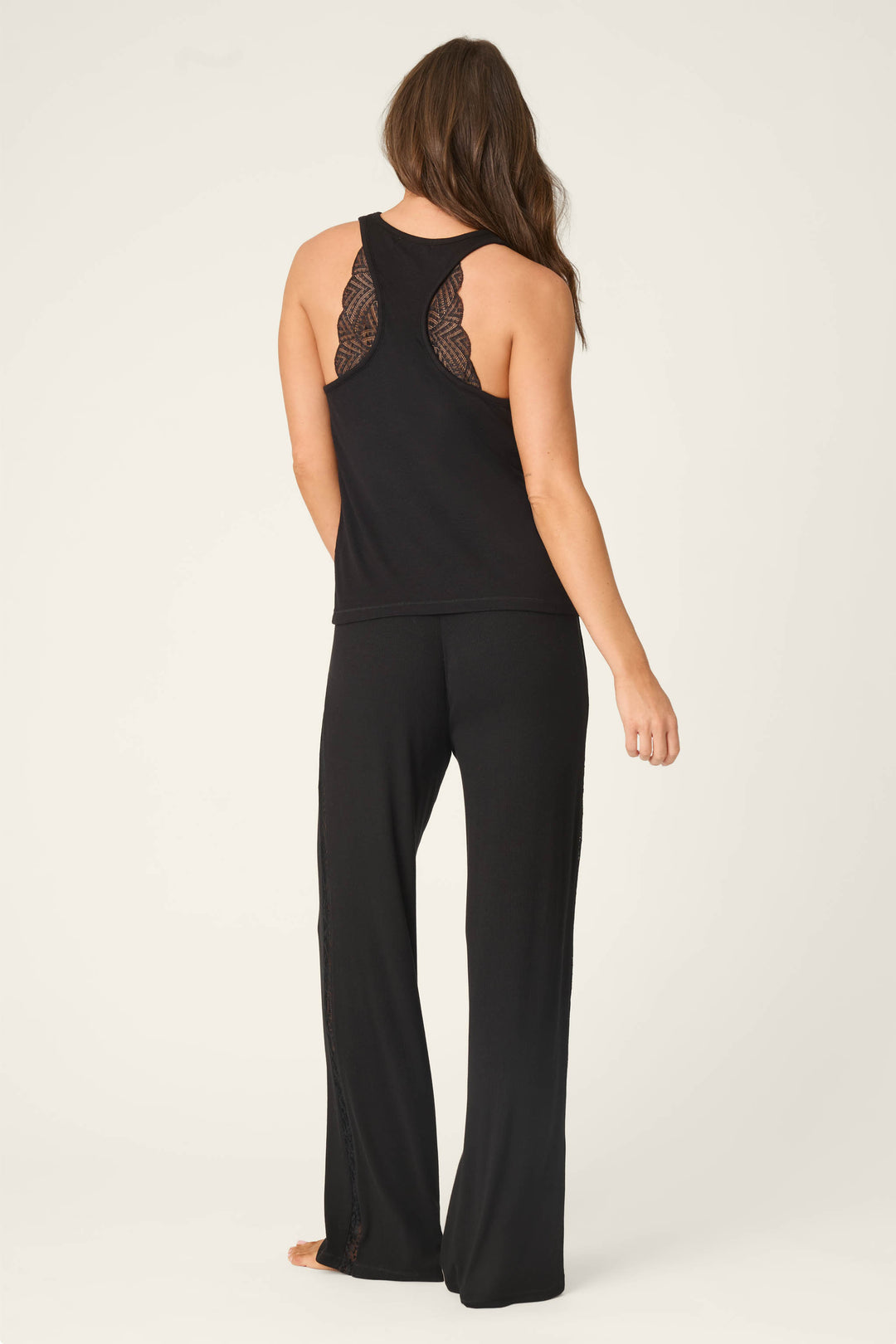 A woman stands barefoot facing away from the camera, wearing the P.J. Salvage Lacey Basics Black Lounge Set against a plain light background.