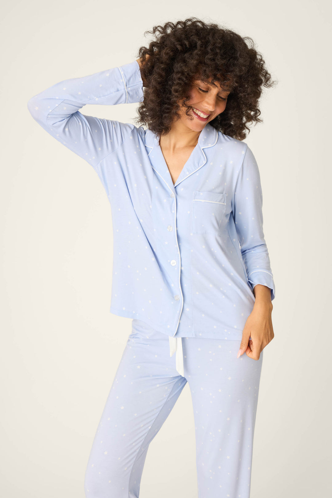 A person with curly hair smiles and poses in the P.J. Salvage Modal Basics Star Print 3-Piece PJ Set, featuring light blue, long-sleeved, button-up pajamas with a subtle white star pattern against a plain background.