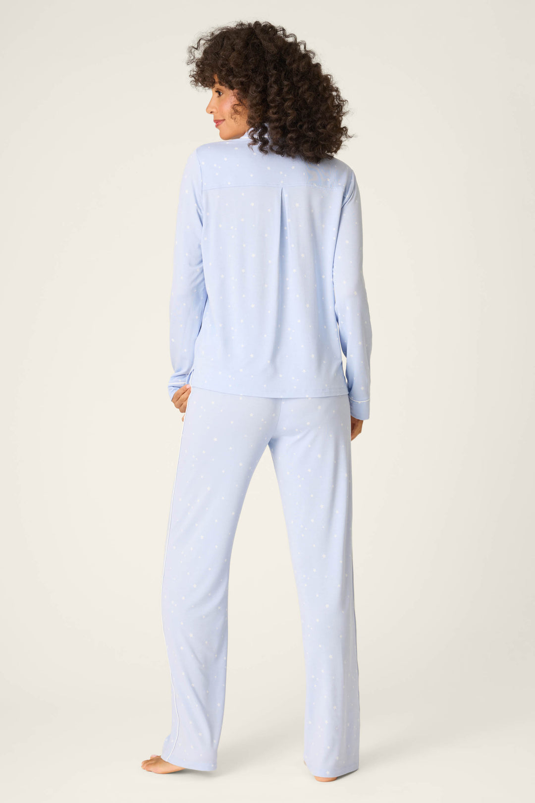 A person with curly hair stands barefoot, back to camera, wearing P.J. Salvage's Modal Basics Star Print 3-Piece PJ Set in light blue on a plain off-white background.