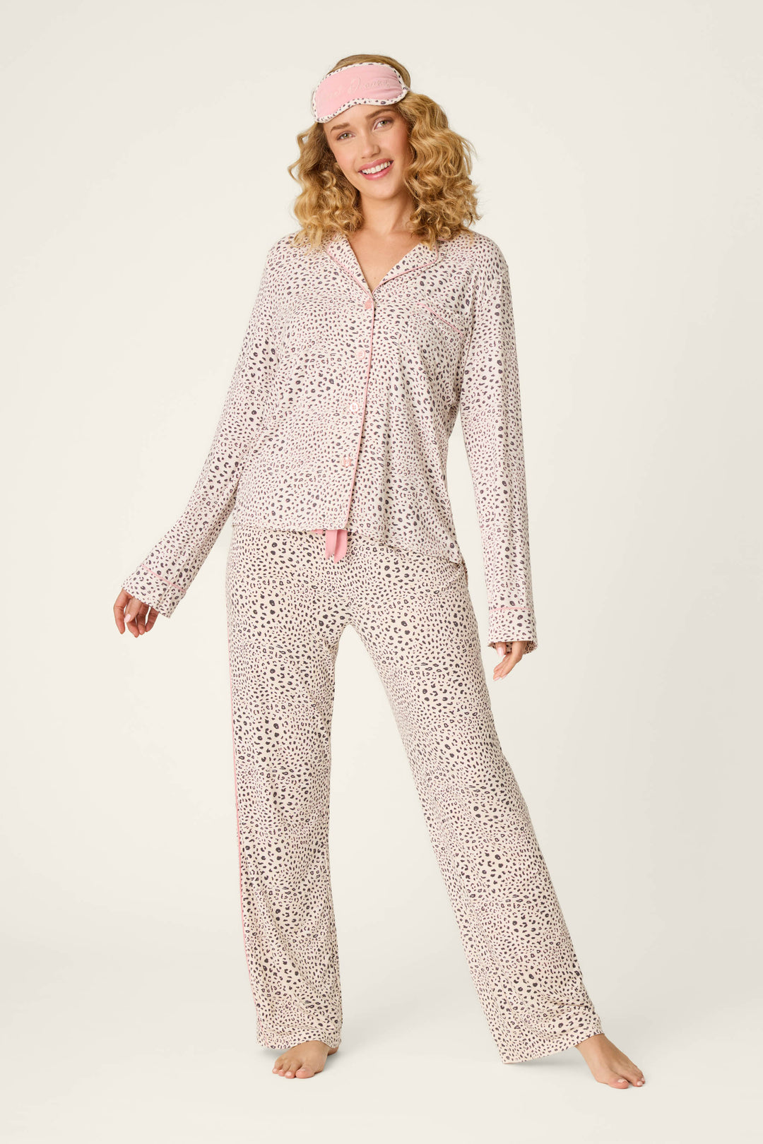 A smiling woman with curly blonde hair models the Modal Dreams Leopard Print 3-Piece PJ Set by P.J. Salvage, featuring a matching sleep mask and standing barefoot against a plain background.