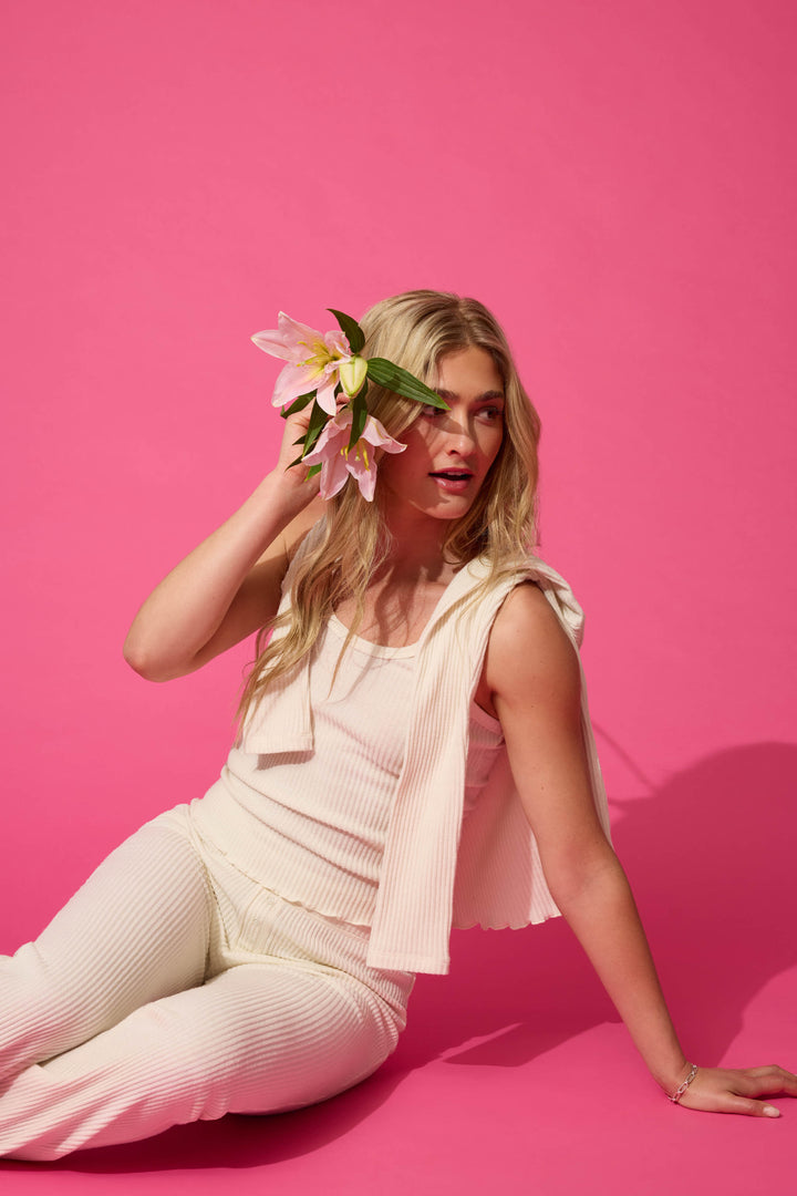 A person with long blonde hair sits on a pink floor against a pink background, holding pink lilies near their head, wearing the P.J. Salvage Reloved Rib Natural 3-Piece Lounge Set.