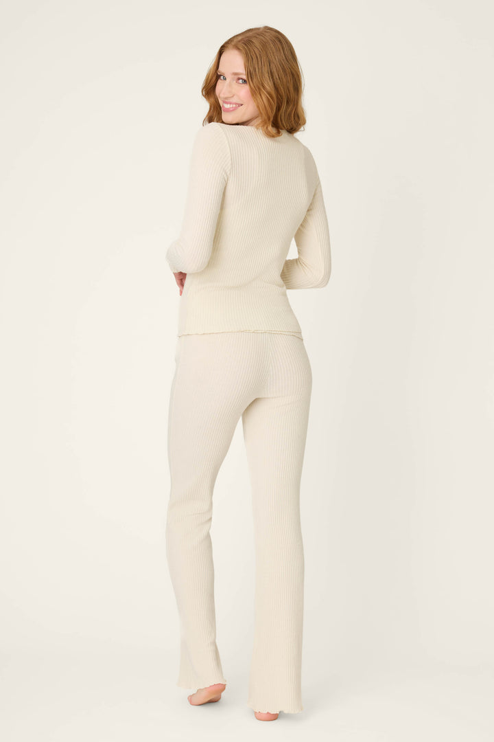 A person with long, wavy, auburn hair stands barefoot, facing away from the camera in the P.J. Salvage Reloved Rib Natural 3-Piece Lounge Set made with REPREVE yarn, against an off-white background.