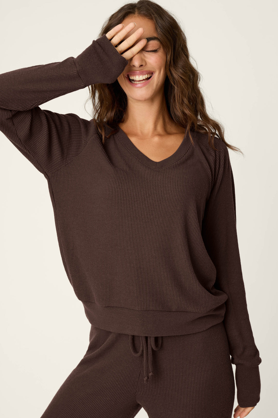 A woman with wavy hair stands against a plain background, smiling as she covers her eyes with her hand. She is wearing the P.J. Salvage Textured Essentials Chocolate Brown Long Sleeve Top.