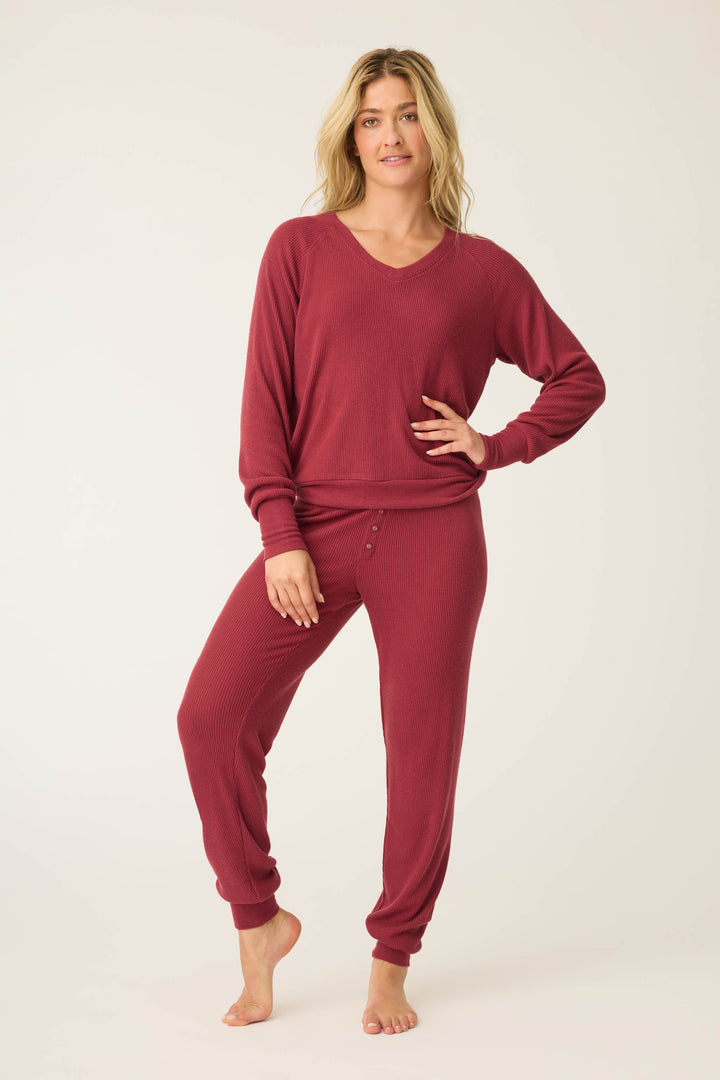 A person stands barefoot against a plain background, wearing the P.J. Salvage Textured Essentials Long Sleeve Top in Port and matching jogger-style pants with waist buttons, posing with one hand on their hip.