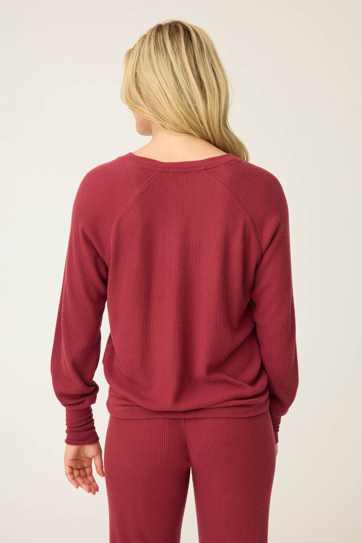 A person with long blonde hair stands with their back to the camera, wearing the P.J. Salvage Textured Essentials Long Sleeve Top in Port and matching ribbed pants.