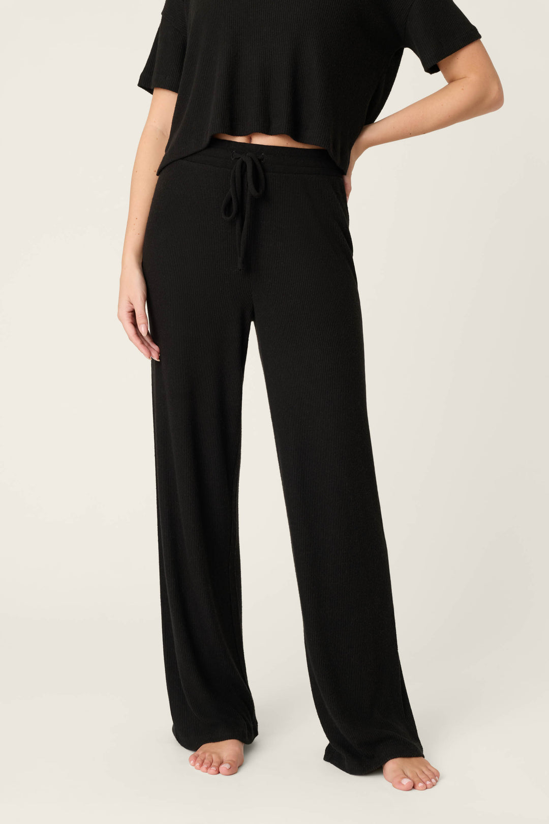 A person wearing P.J. Salvage Textured Essentials Black Open Leg Pants with a drawstring waistband and a black short-sleeve top stands barefoot against a plain light background.