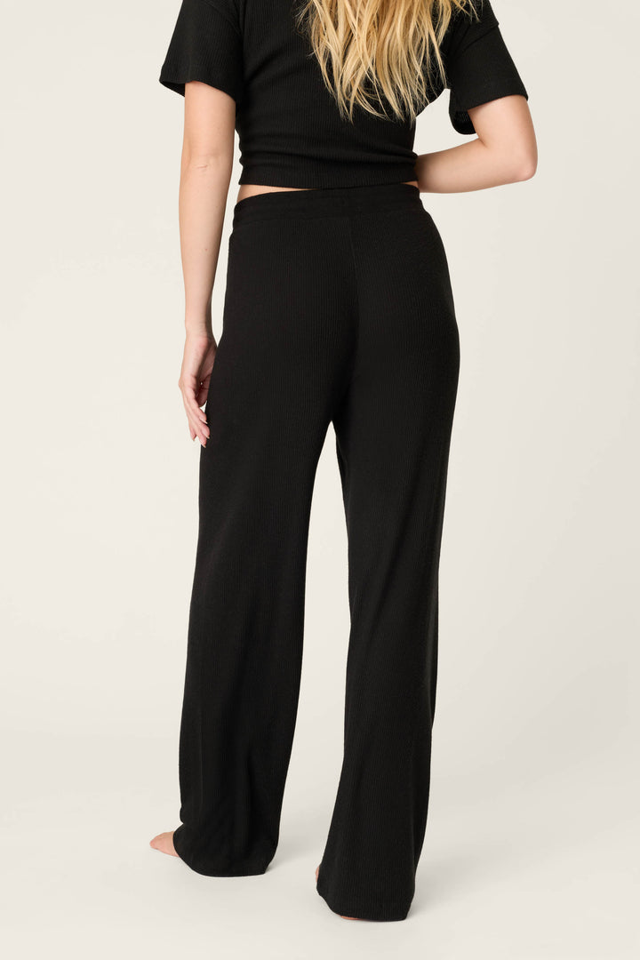 A person with long blonde hair stands barefoot on a light backdrop, wearing the P.J. Salvage Textured Essentials Black Open Leg Pants and a short-sleeve black top, viewed from behind.