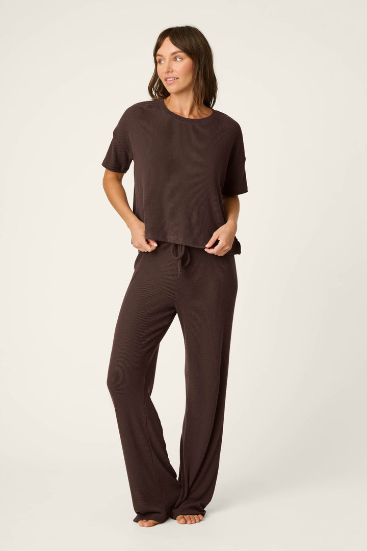 A woman stands barefoot, wearing the P.J. Salvage Textured Essentials Chocolate Brown Lounge Pants with a matching loose-fitting short-sleeve top. She looks to the side with one hand on her hip against a plain, light background.