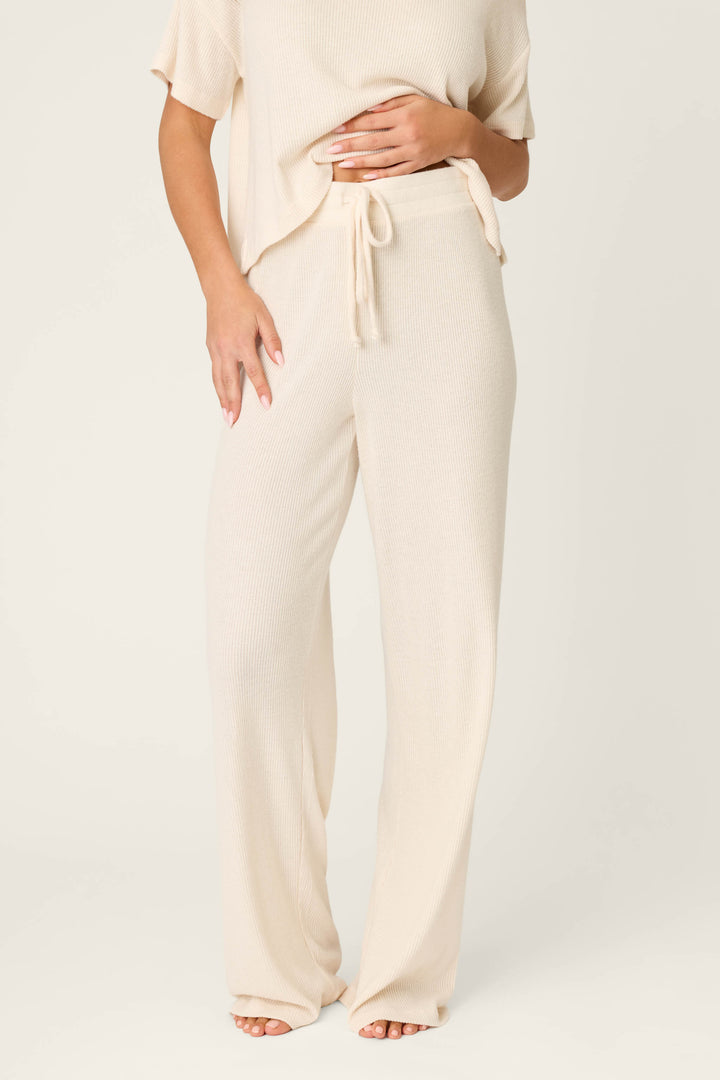 A person wears P.J. Salvage's Textured Essentials Stone Open Leg Pants—cream-colored, ribbed knit with a drawstring waist—and a matching short-sleeve top, standing barefoot against a plain light background.