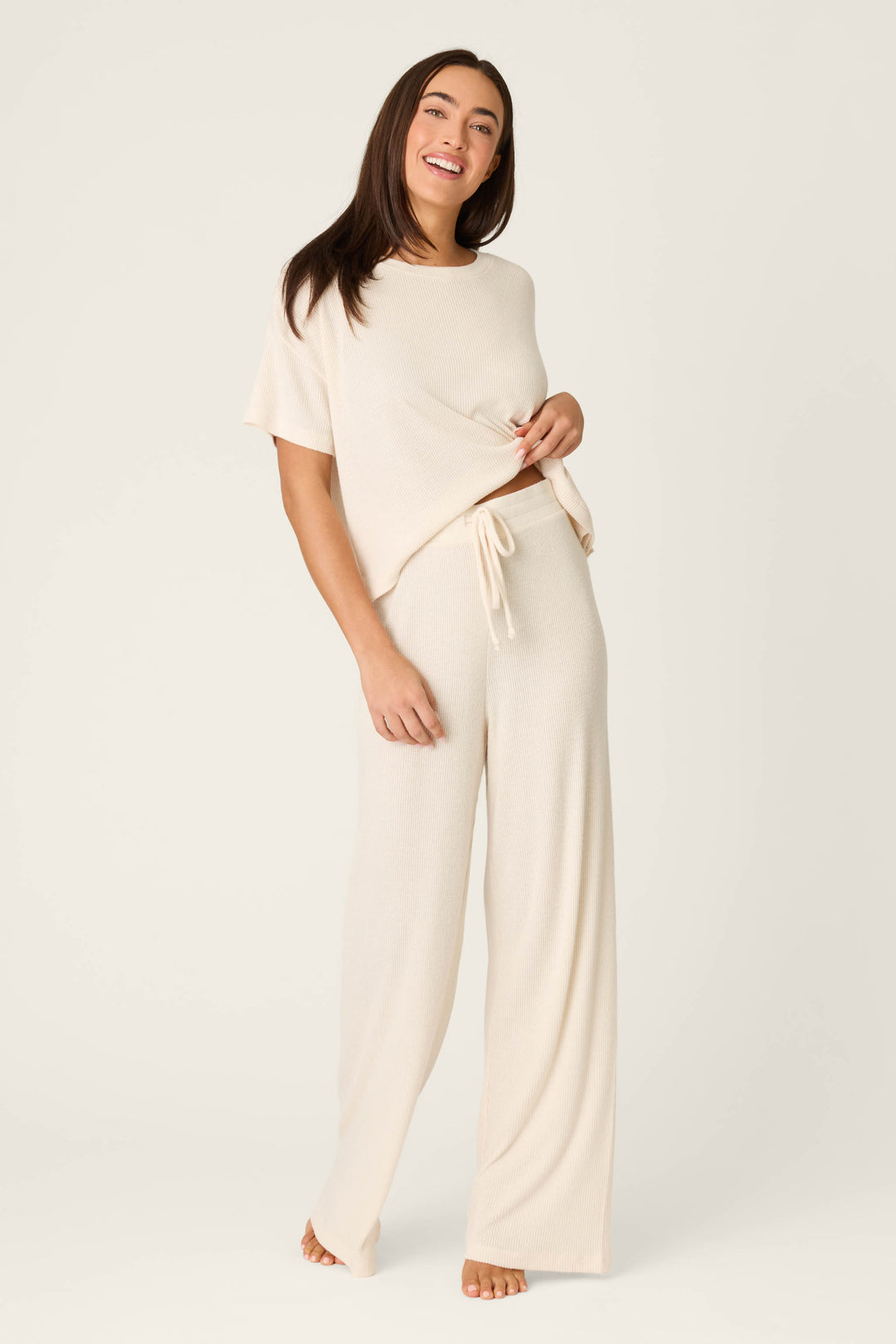 A woman stands barefoot against a plain background, smiling and wearing P.J. Salvage’s Textured Essentials Stone Open Leg Pants with a matching cream short-sleeve top, holding part of her top in one hand.