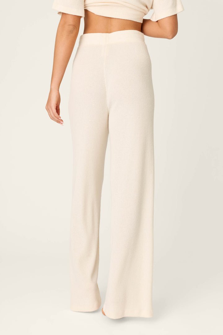 A person stands barefoot on a neutral background, seen from the back, wearing P.J. Salvage Textured Essentials Stone Open Leg Pants paired with a matching cream ribbed cropped short-sleeve top.