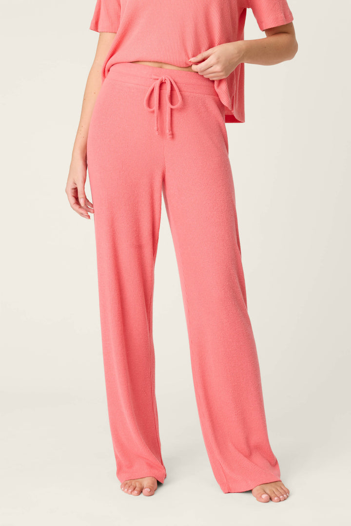 A person models P.J. Salvage’s Textured Essentials Sunset Rose Open Leg Pants—loose, high-waisted, pink ribbed with a drawstring—paired with a matching short-sleeve top, barefoot against a plain background.