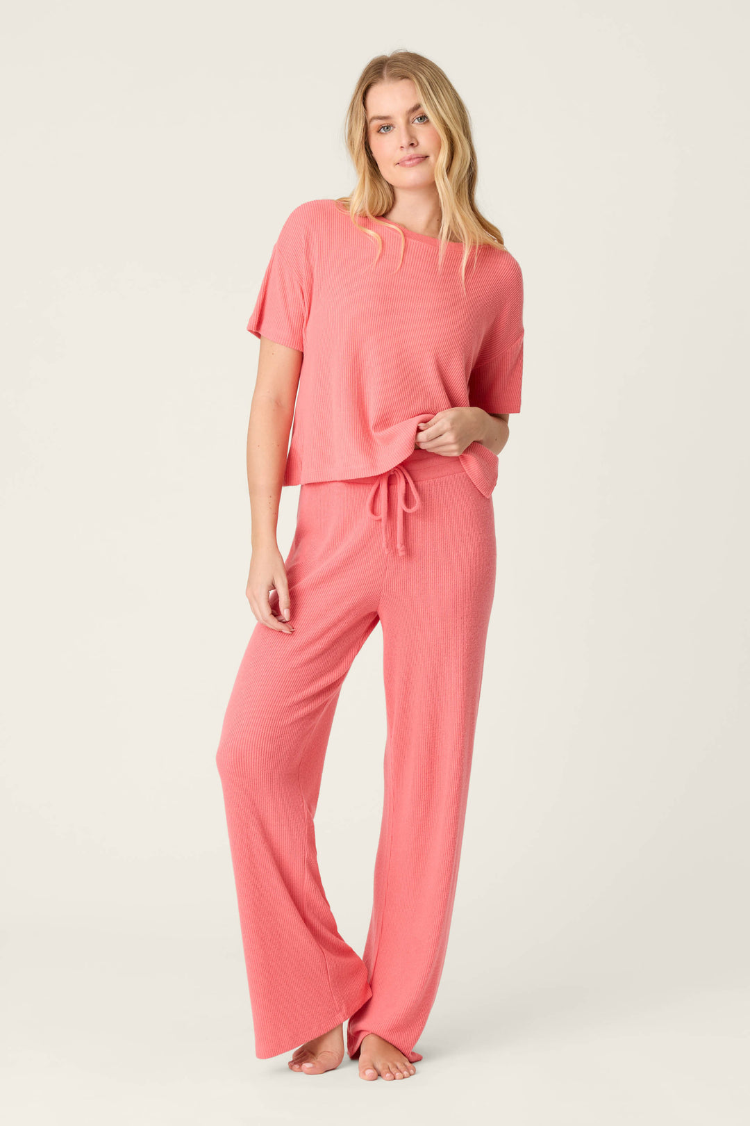 A person stands barefoot against a plain light background, wearing P.J. Salvage’s Textured Essentials Sunset Rose Open Leg Pants with a matching pink short-sleeve top. One hand is in their pocket and they appear relaxed.