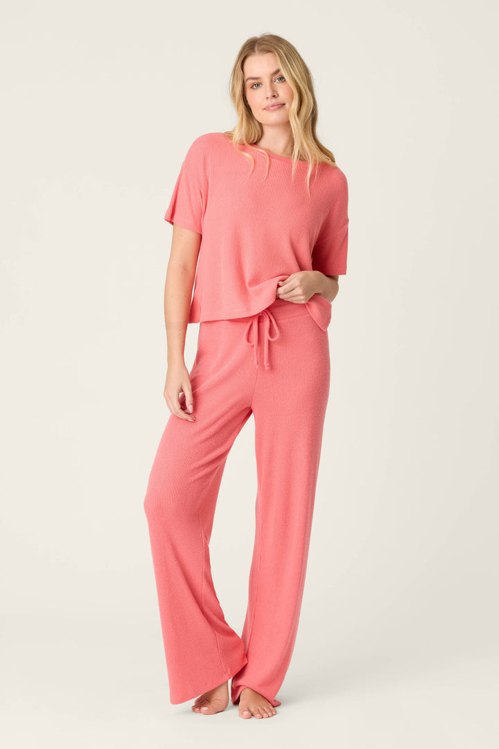 A person stands barefoot against a plain light background, wearing P.J. Salvage’s Textured Essentials Sunset Rose Open Leg Pants with a matching pink short-sleeve top. One hand is in their pocket and they appear relaxed.