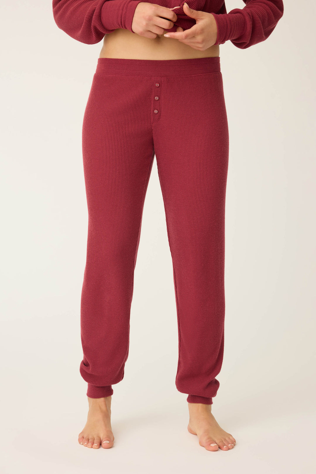 A person stands barefoot in P.J. Salvage Textured Essentials Banded Pants - Port, shown from waist to ankles against a plain background. The burgundy pants feature ribbed texture and a buttoned front.