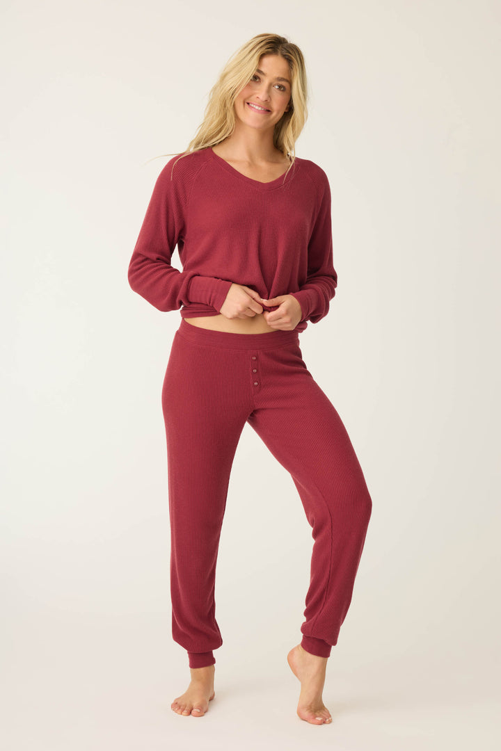 A woman stands barefoot against a light background, smiling as she holds her shirt at the waist while wearing P.J. Salvage’s Textured Essentials Banded Pants in Port—a matching long-sleeve burgundy pajama set featuring button details on the pants.