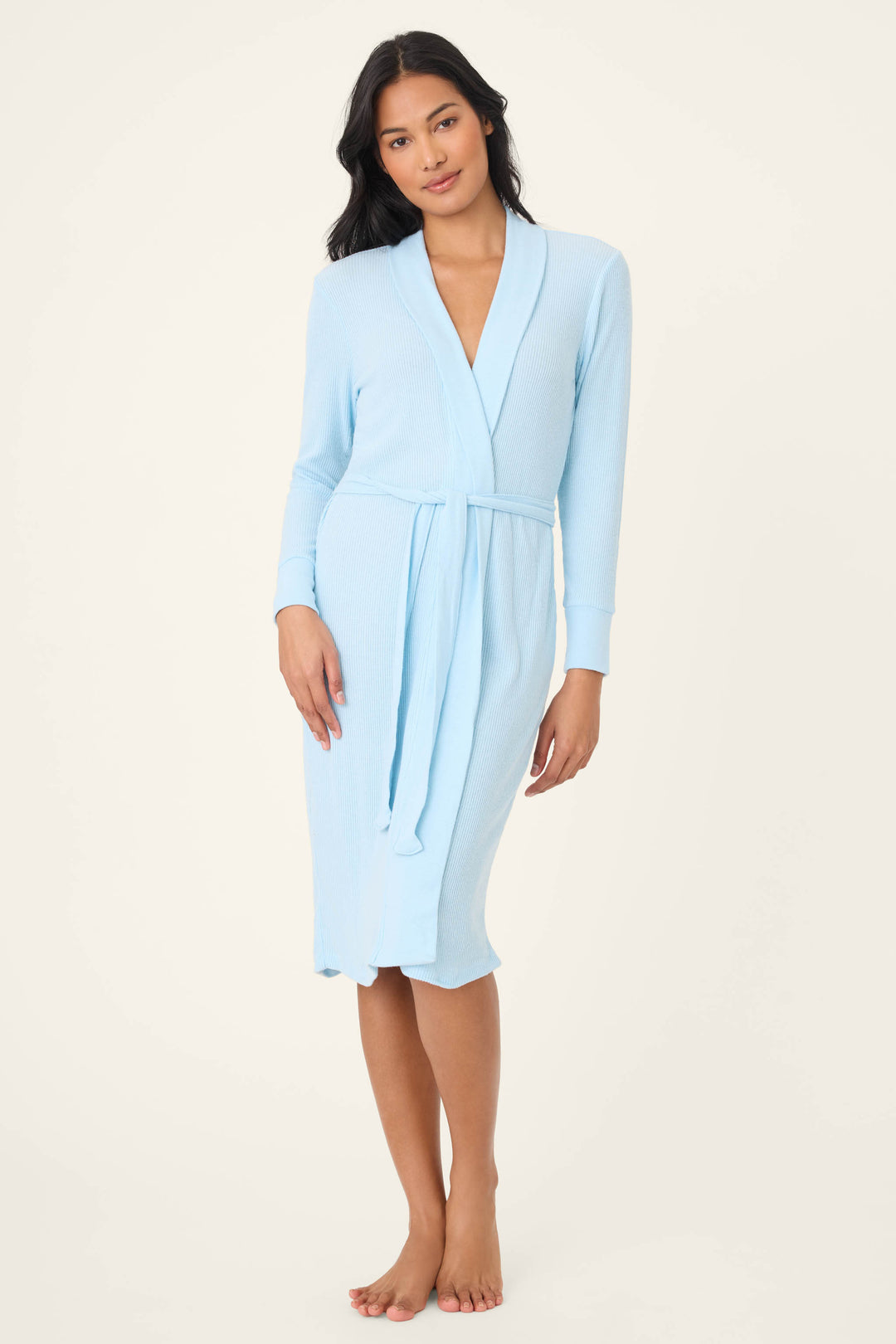 A woman stands barefoot in the P.J. Salvage Textured Essentials Robe, a sky-blue, ribbed cotton-blend jersey lounge robe tied at the waist. She has long, dark hair and is set against a plain, light background.
