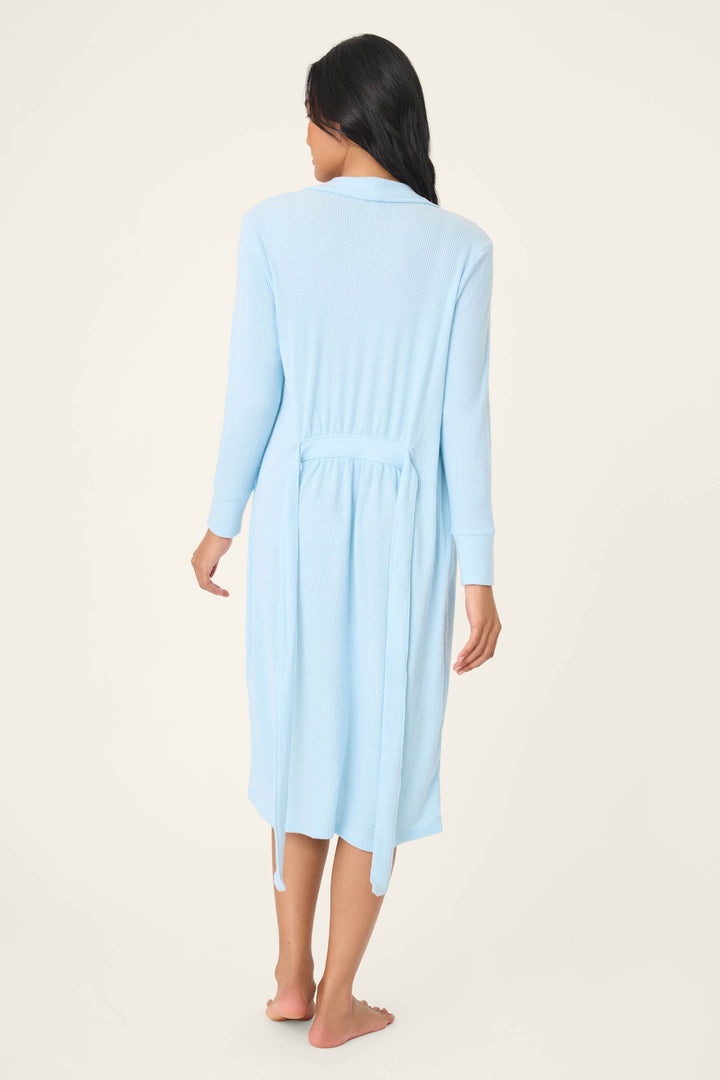 A person with long dark hair stands barefoot facing away, wearing the P.J. Salvage Textured Essentials Robe in sky blue. The ribbed, knee-length style with a back-tied belt stands out against the plain background, highlighting its cozy appeal.