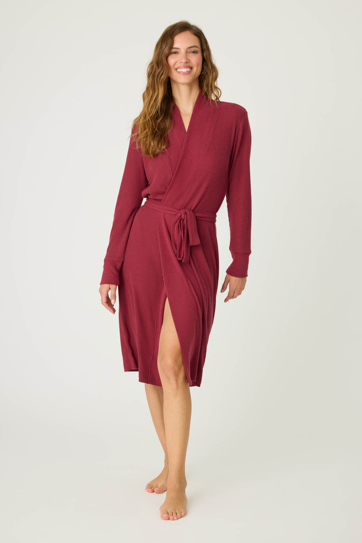 A woman with long, wavy hair wears the P.J. Salvage Textured Essentials Robe in Port, a knee-length, long-sleeved maroon robe with a waist tie. Barefoot and smiling slightly, she stands against a plain light background.
