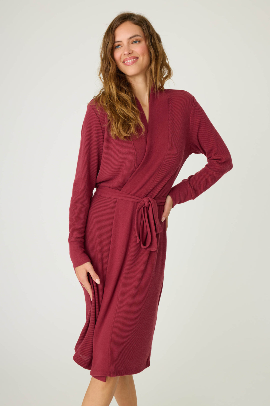 A woman stands against a plain background wearing the P.J. Salvage Textured Essentials Robe in Port. The long-sleeved, knee-length robe features a tied waist belt. She smiles, with one hand on her hip and the other resting by her side.
