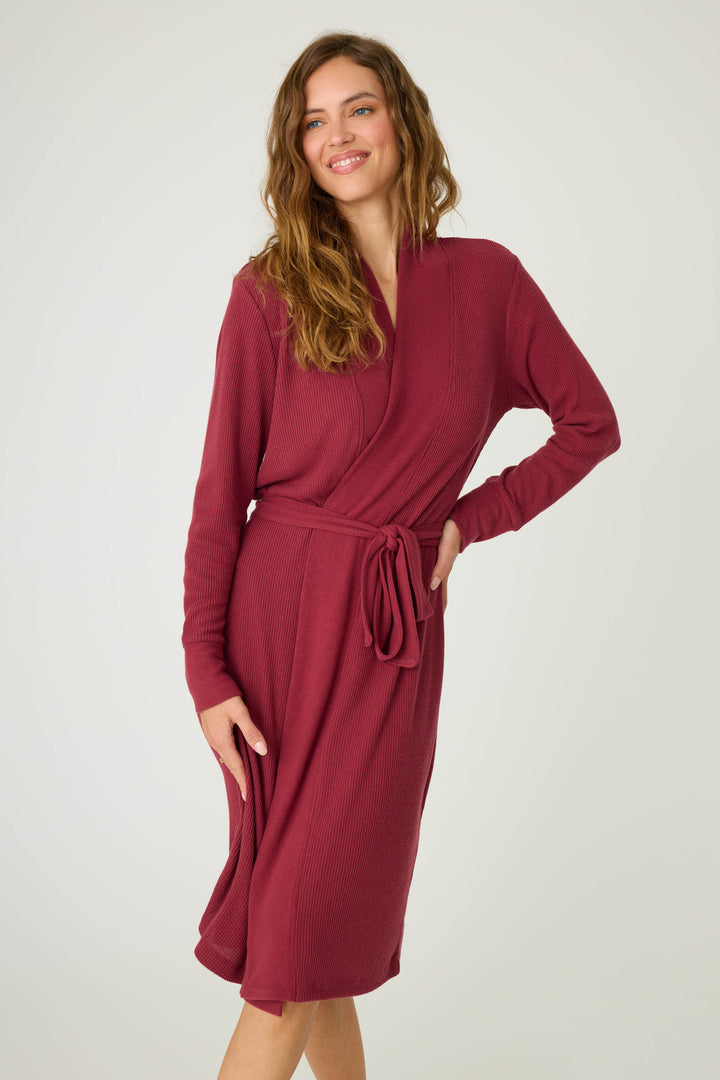 A woman stands against a plain background wearing the P.J. Salvage Textured Essentials Robe in Port. The long-sleeved, knee-length robe features a tied waist belt. She smiles, with one hand on her hip and the other resting by her side.