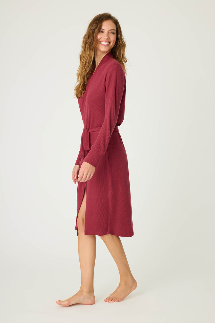 A woman with long hair stands barefoot, smiling and looking to the side, wearing the knee-length, long-sleeve P.J. Salvage Textured Essentials Robe in Port with a matching belt against a plain light background.