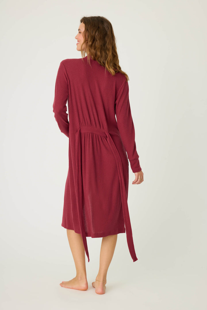 A woman stands barefoot facing away, wearing the P.J. Salvage Textured Essentials Robe in Port—a long-sleeved, knee-length robe with a loosely tied belt. The background is plain and light-colored.