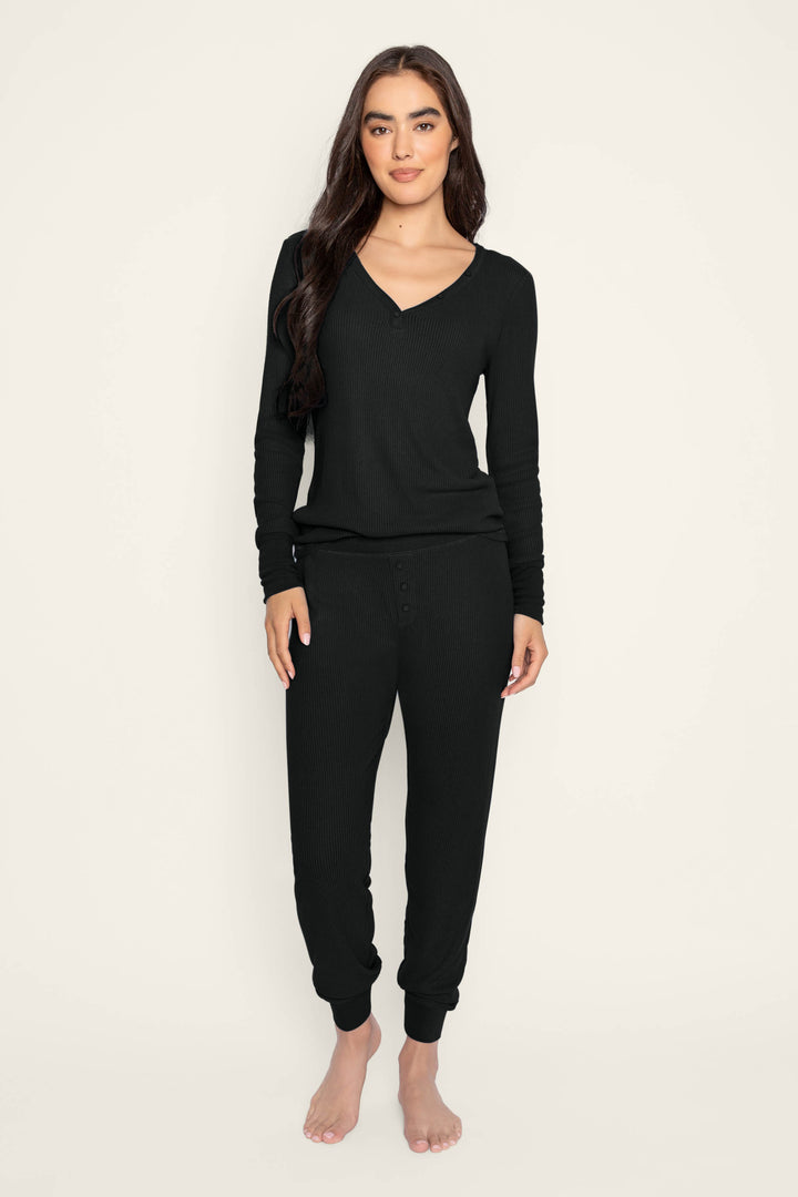 A woman with long dark hair stands barefoot against a plain background, wearing the P.J. Salvage Textured Essentials Black Lounge Set—a matching ribbed ensemble featuring a relaxed V-neck long-sleeve top and jogger-style pants. She faces forward with a relaxed expression.