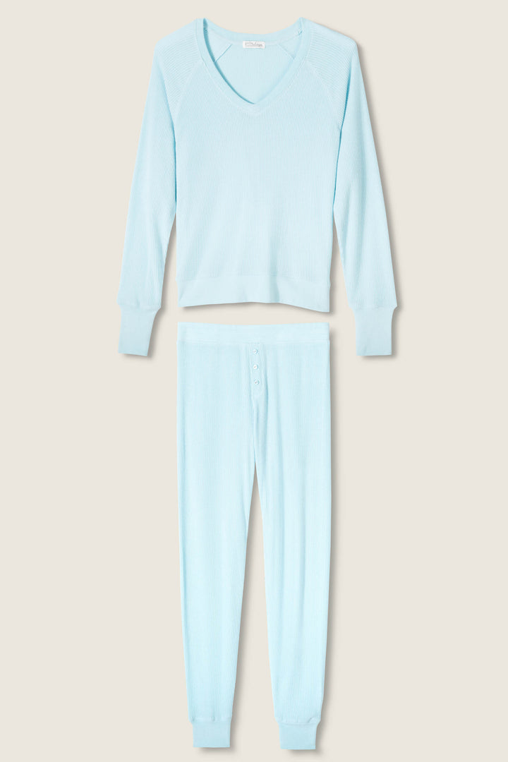 The P.J. Salvage Textured Essentials Light Sky Pants Set features a light blue ribbed V-neck long-sleeve pajama top and matching pants with button fly and cuffed ankles, shown on a neutral background.