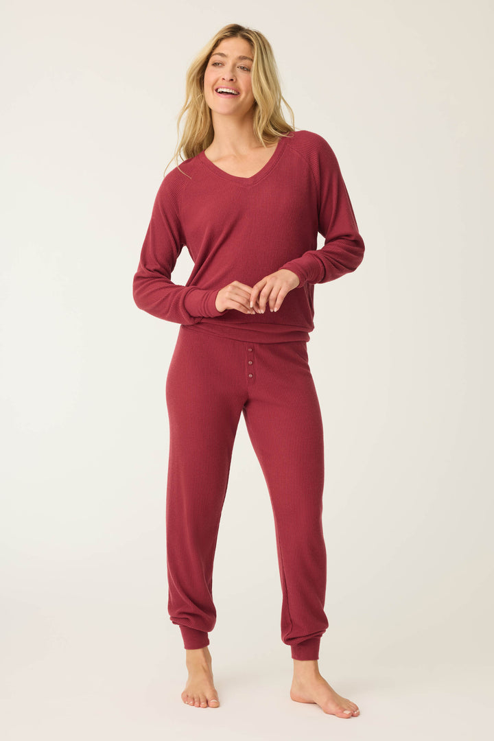 A woman, barefoot on a light background, wears the P.J. Salvage Textured Essentials PJ Set— a matching long-sleeve burgundy pajama with button-front pants. She smiles and holds her waistband with both hands.